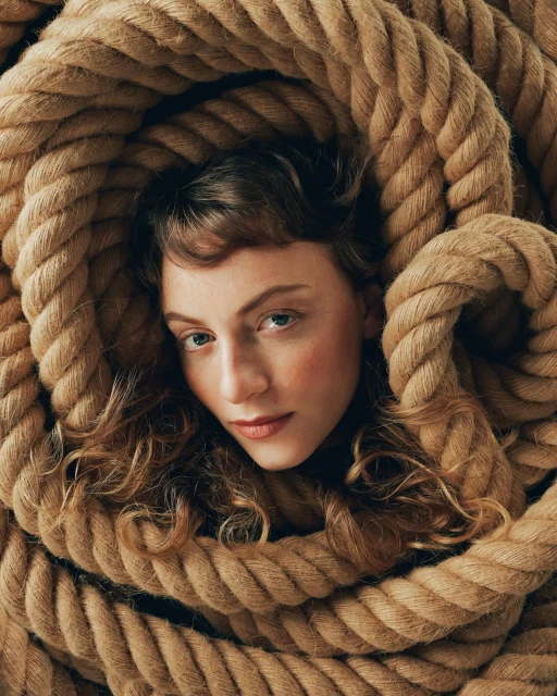 Eye-level portrait of an adult woman with curly hair, soft makeup, and neutral expression, framed by thick coils of tan rope.
