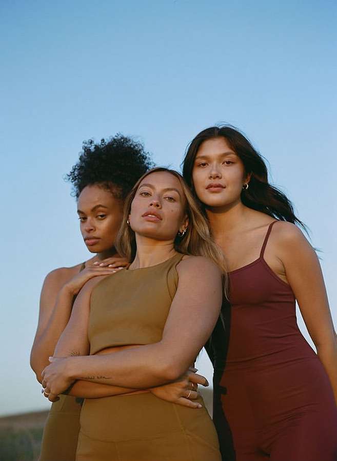 Three diverse women in fitness apparel pose outdoors. One woman in olive green crosses arms, another in red straps are visible, blue sky.