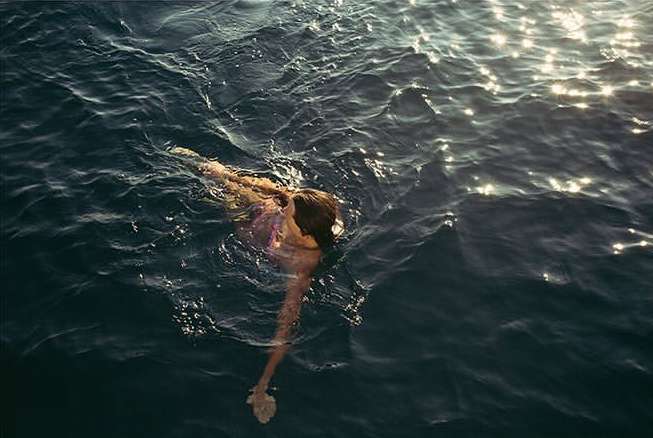 Woman swimming in the dark blue ocean water with sunlight reflecting off the surface in a serene outdoor setting, aerial view.