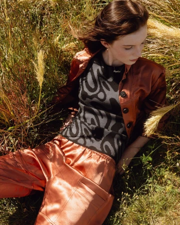 Young woman in a glossy burnt orange jacket and satin trousers lying in a sunlit tall grass meadow.