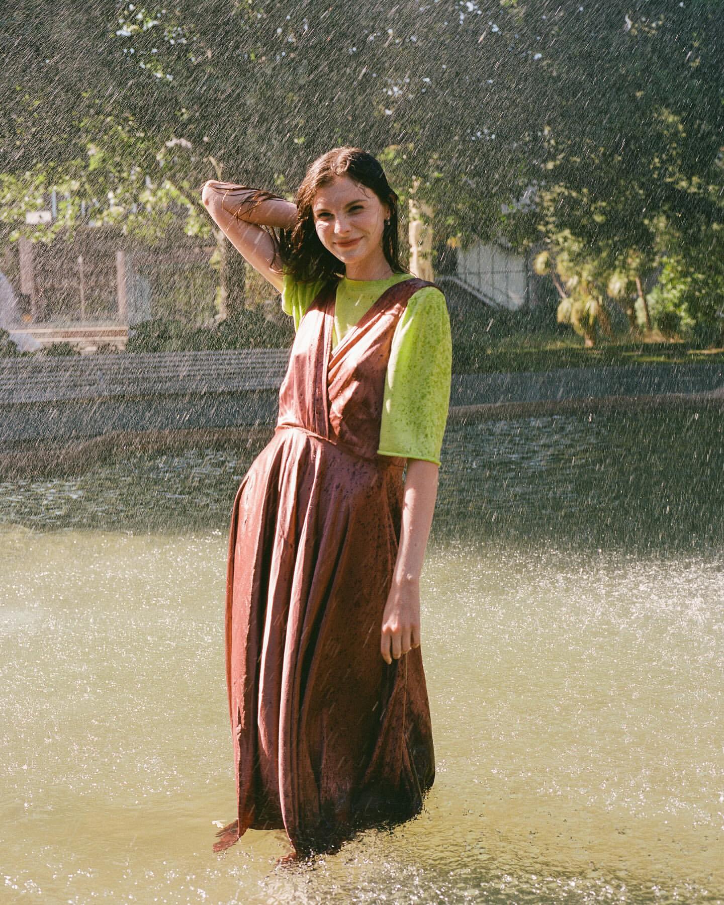 Woman in a brown dress and green shirt standing in water while heavy rain falls around her.