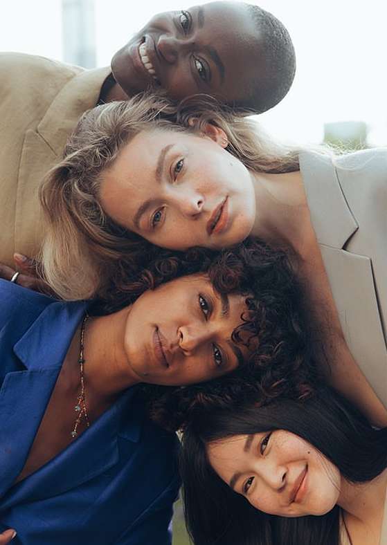 Portrait of four diverse women laying their heads together in a studio setting. They wear fashionable blazers in different colors.