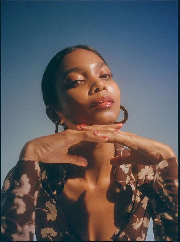 Headshot of a young Black female model wearing a brown floral dress, hoop earrings, and red nails against a blue sky background.