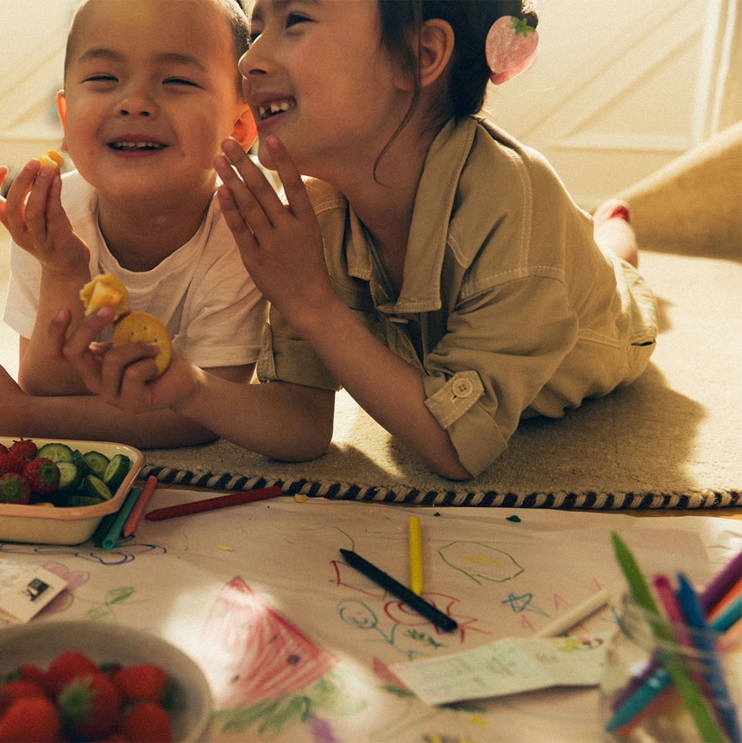 Two smiling Asian children share snacks while lying on a rug and drawing with crayons, in a brightly lit indoor setting, daytime.