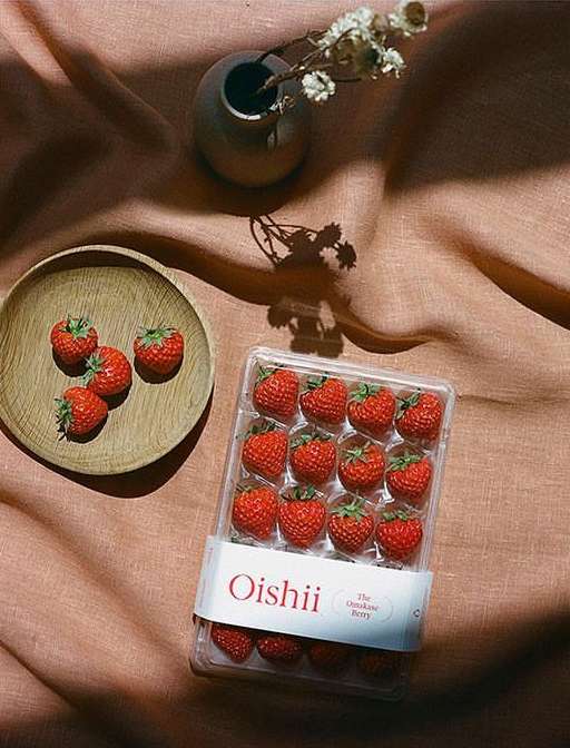 Overhead shot displays strawberries in a wooden dish and plastic container, with a vase of flowers set against a pink fabric.