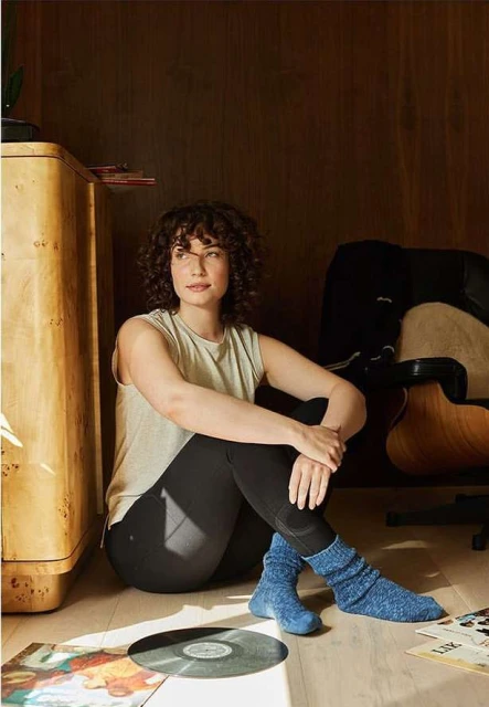 Young woman in socks sitting on the floor with vinyl records next to a cabinet and chair in a home interior with dark wood walls.