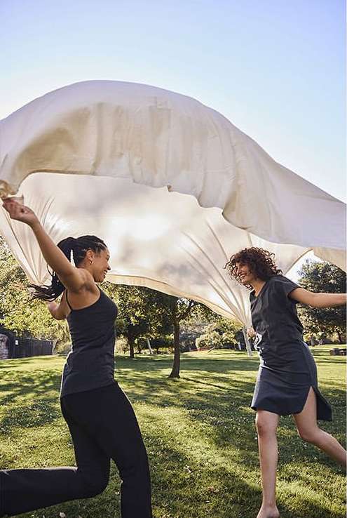 Two adult women laugh and hold a white cloth overhead in a sunny grassy park setting with trees and blue sky as a backdrop.