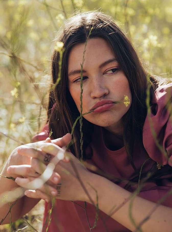 Portrait of a young woman with long brown hair posing in a field of yellow wildflowers, wearing a pink top and looking contemplative.