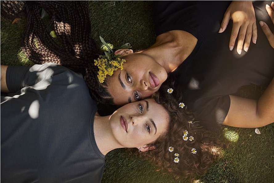 Two women lie head-to-head on green grass. One with braids and yellow flowers in hair, the other with curls and white flowers.