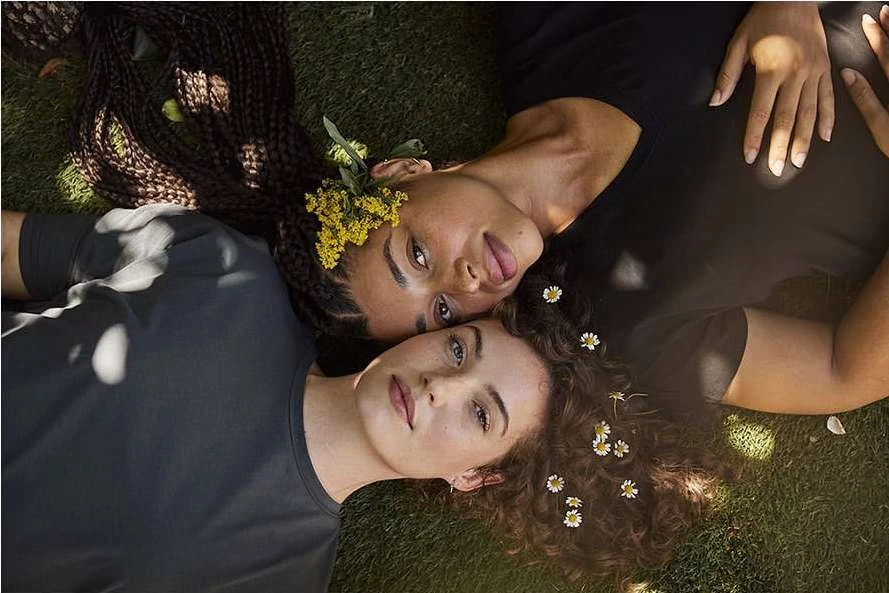 Two women lie head-to-head on green grass. One with braids and yellow flowers in hair, the other with curls and white flowers.