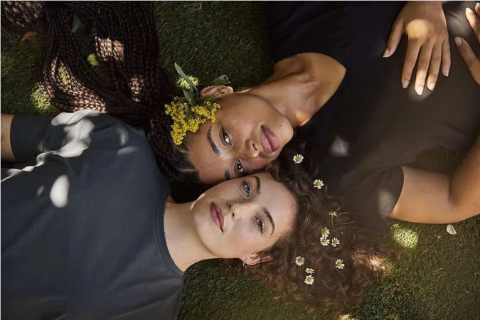 Two women lie head-to-head on green grass. One with braids and yellow flowers in hair, the other with curls and white flowers.