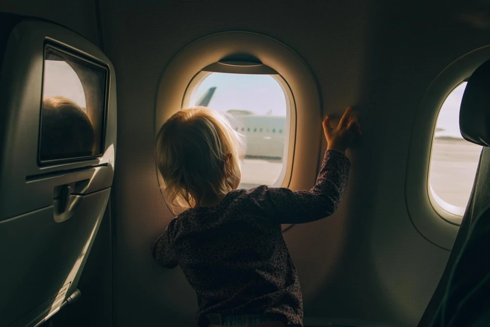 Child looks out of an airplane window at the wing and airport; bright sunshine creates a serene travel scene. Airplane interior.