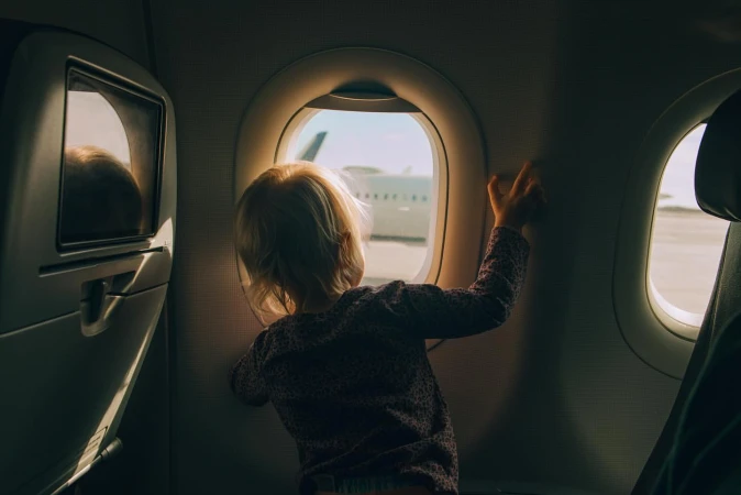 Child looks out of an airplane window at the wing and airport; bright sunshine creates a serene travel scene. Airplane interior.