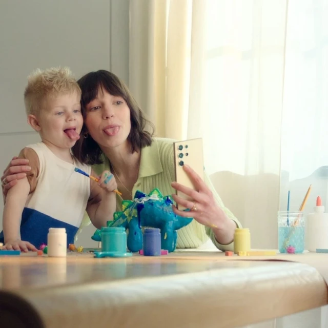 Mother and child taking a selfie while painting a blue dinosaur figurine at a table with art supplies in a brightly lit room.