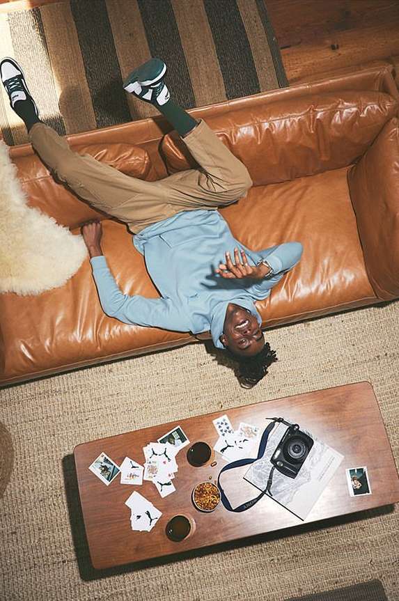 Young man laying upside down on a leather couch with a table in the foreground. The table has playing cards, a camera, and a snack.