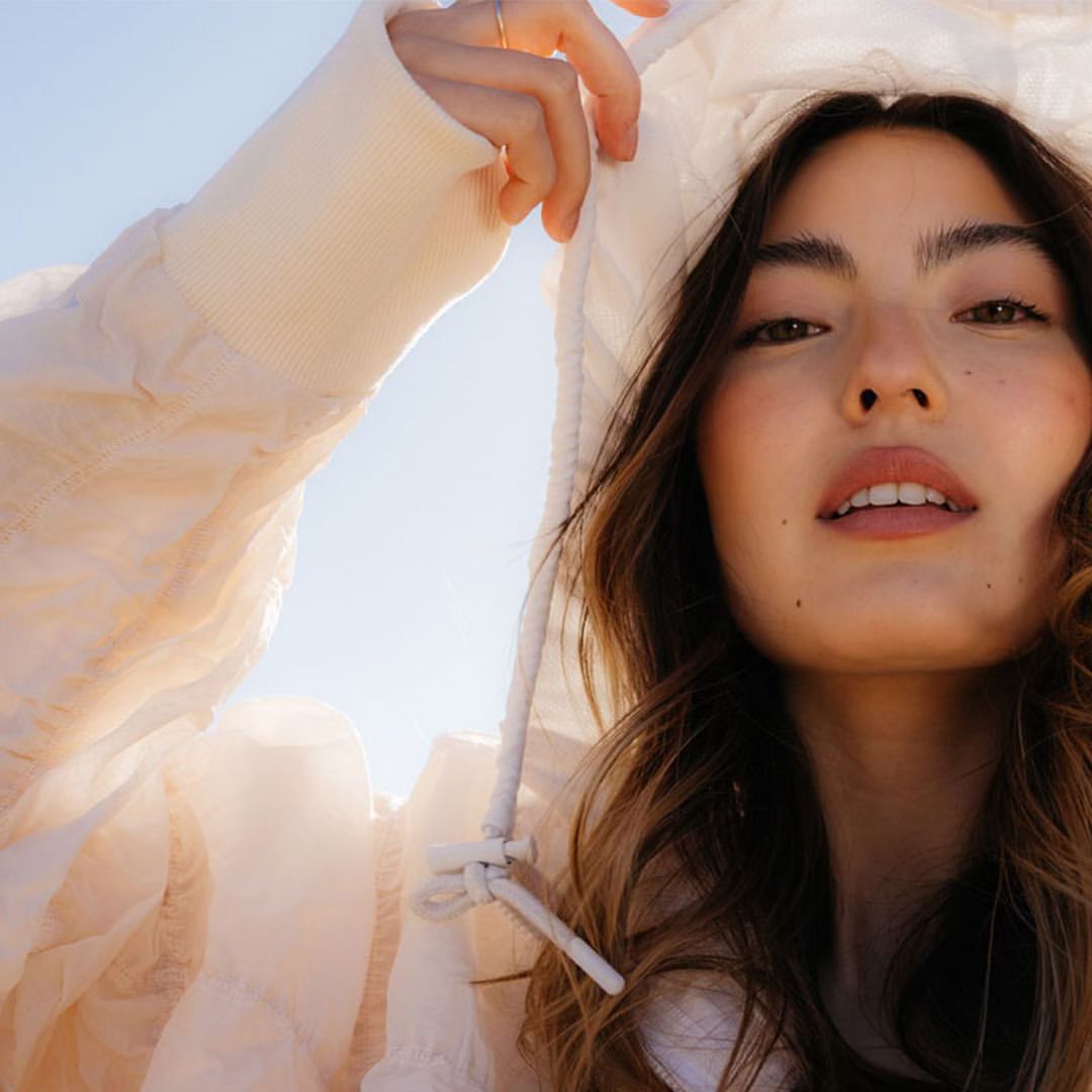 Woman wearing a white hooded jacket looks at the camera outdoors against a blue sky backdrop on a bright, sunny day.