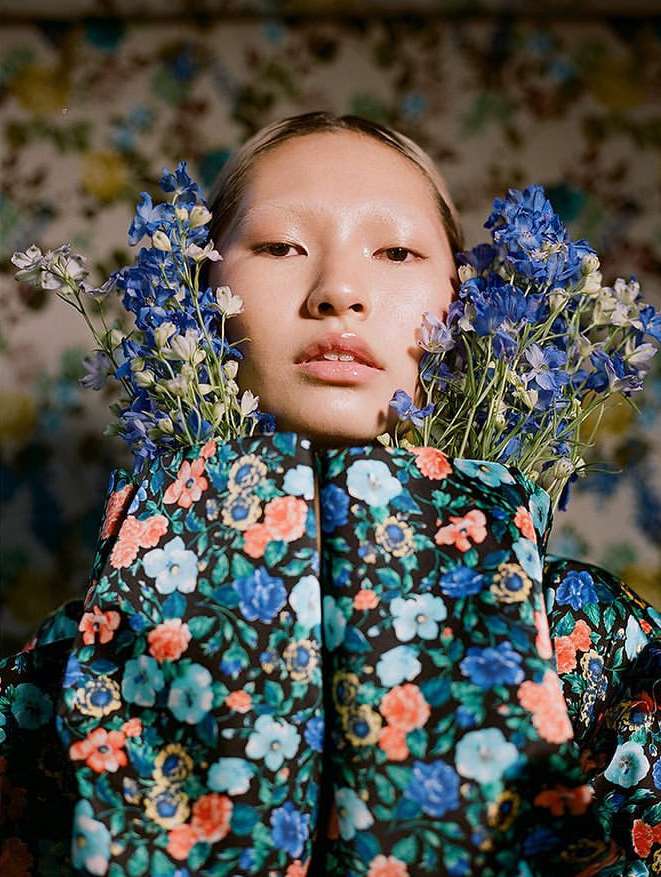 Portrait of an Asian woman wearing a floral patterned jacket with fresh blue flowers around her face, set against a floral wallpaper.