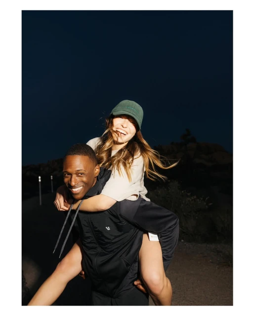 Smiling man giving a piggyback ride to a smiling woman wearing a hat on a path outdoors with dark blue sky and rock formations.
