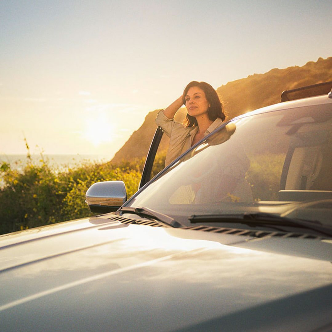 Woman in tan shirt leans against silver SUV parked near a coastline at sunset. The SUV's hood and windshield are in the foreground.