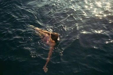 High angle shot of a woman swimming in dark water with glittering sunlight reflections on the surface.