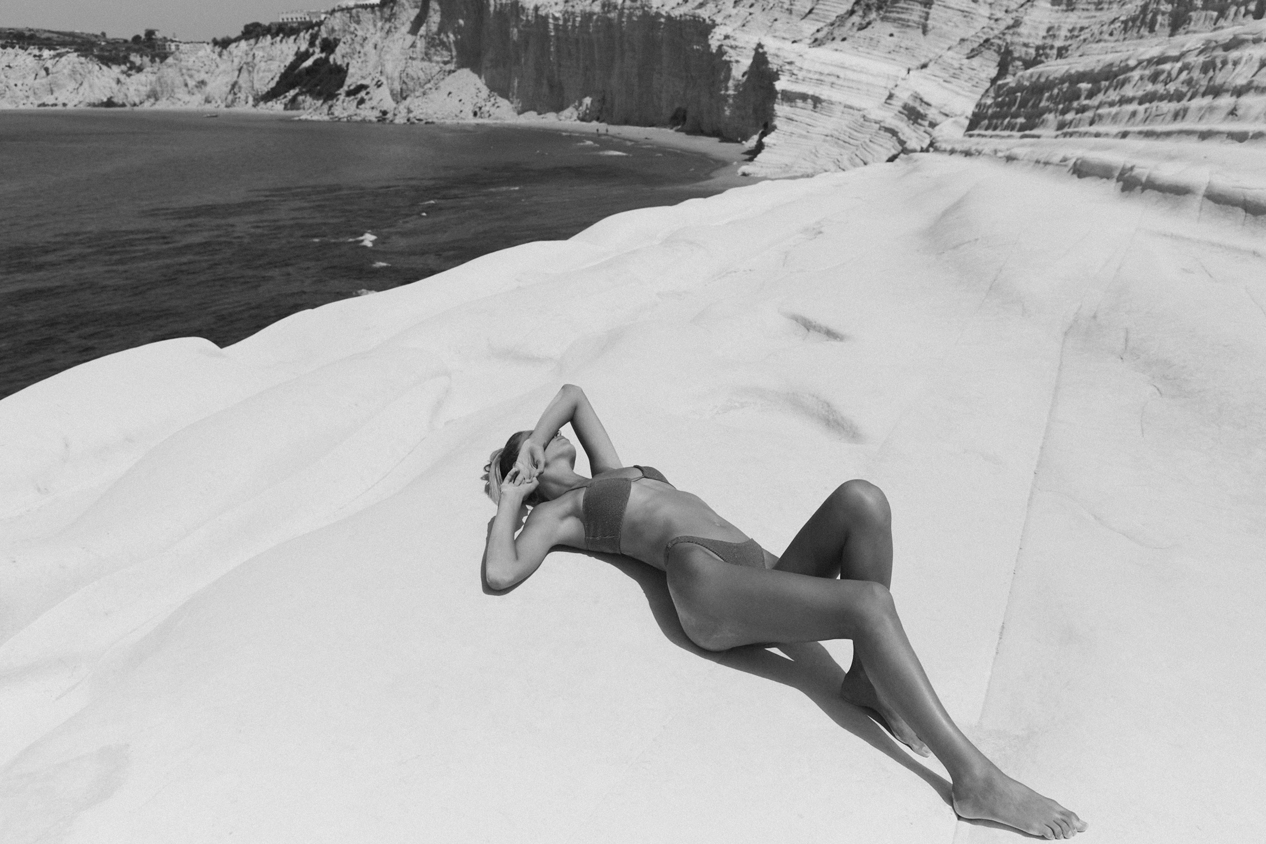 Woman in bikini lying on white rock formation near the ocean with cliffs in the background, enjoying sun and nature.
