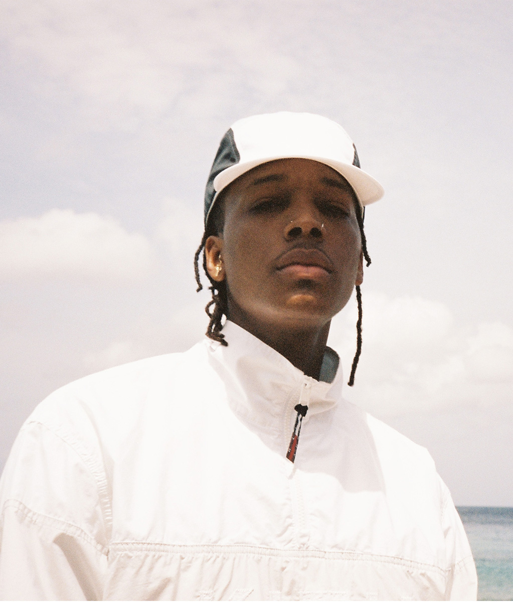 Headshot of a young adult with dark skin and dark hair wearing a white and grey hat and white jacket against a bright, cloudy sky.