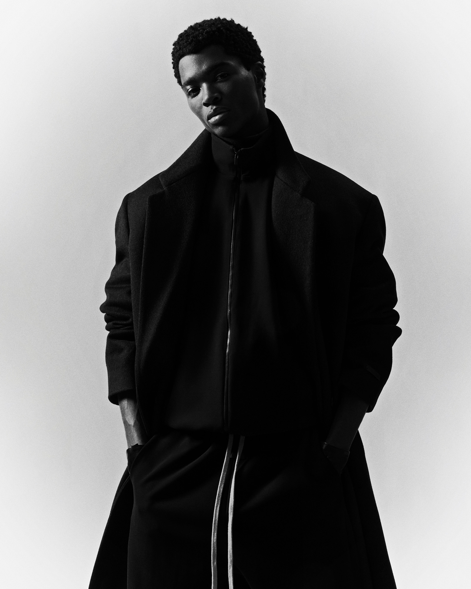 A striking black and white fashion shot of an adult male model with a dark complexion wearing a dark overcoat, turtleneck, and trousers.