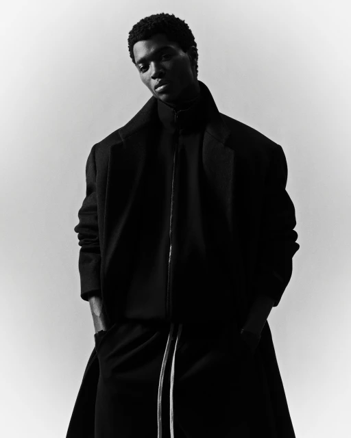 A striking black and white fashion shot of an adult male model with a dark complexion wearing a dark overcoat, turtleneck, and trousers.