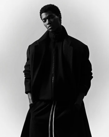 A striking black and white fashion shot of an adult male model with a dark complexion wearing a dark overcoat, turtleneck, and trousers.