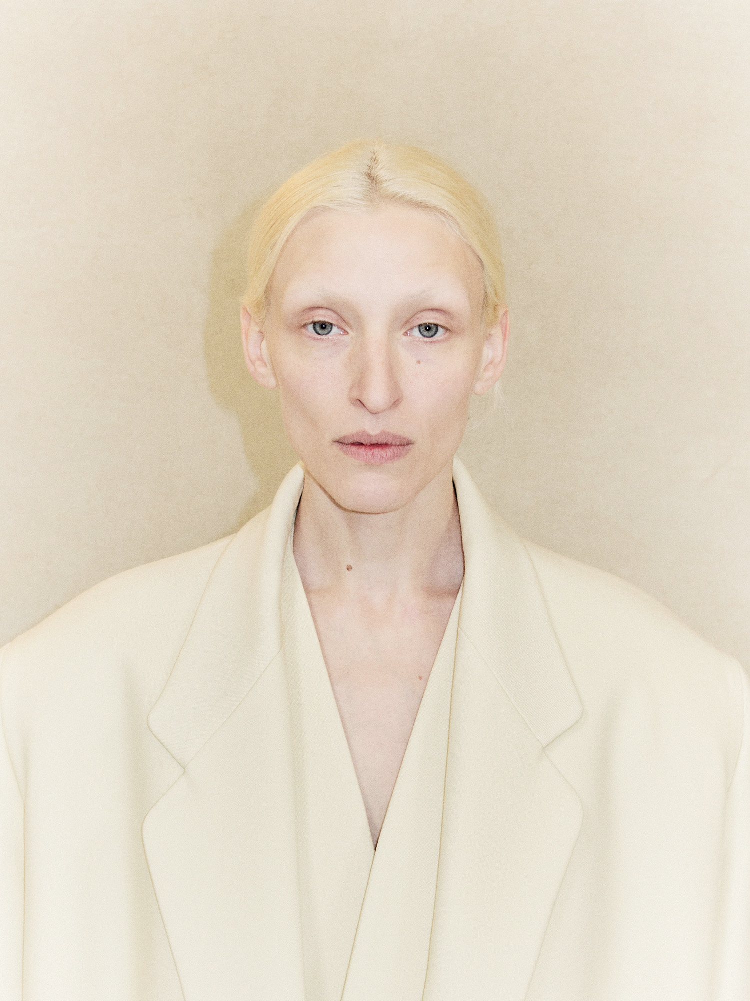 Portrait of an adult woman with fair skin, light blonde hair, wearing a cream-colored blazer, posing against a neutral beige background.