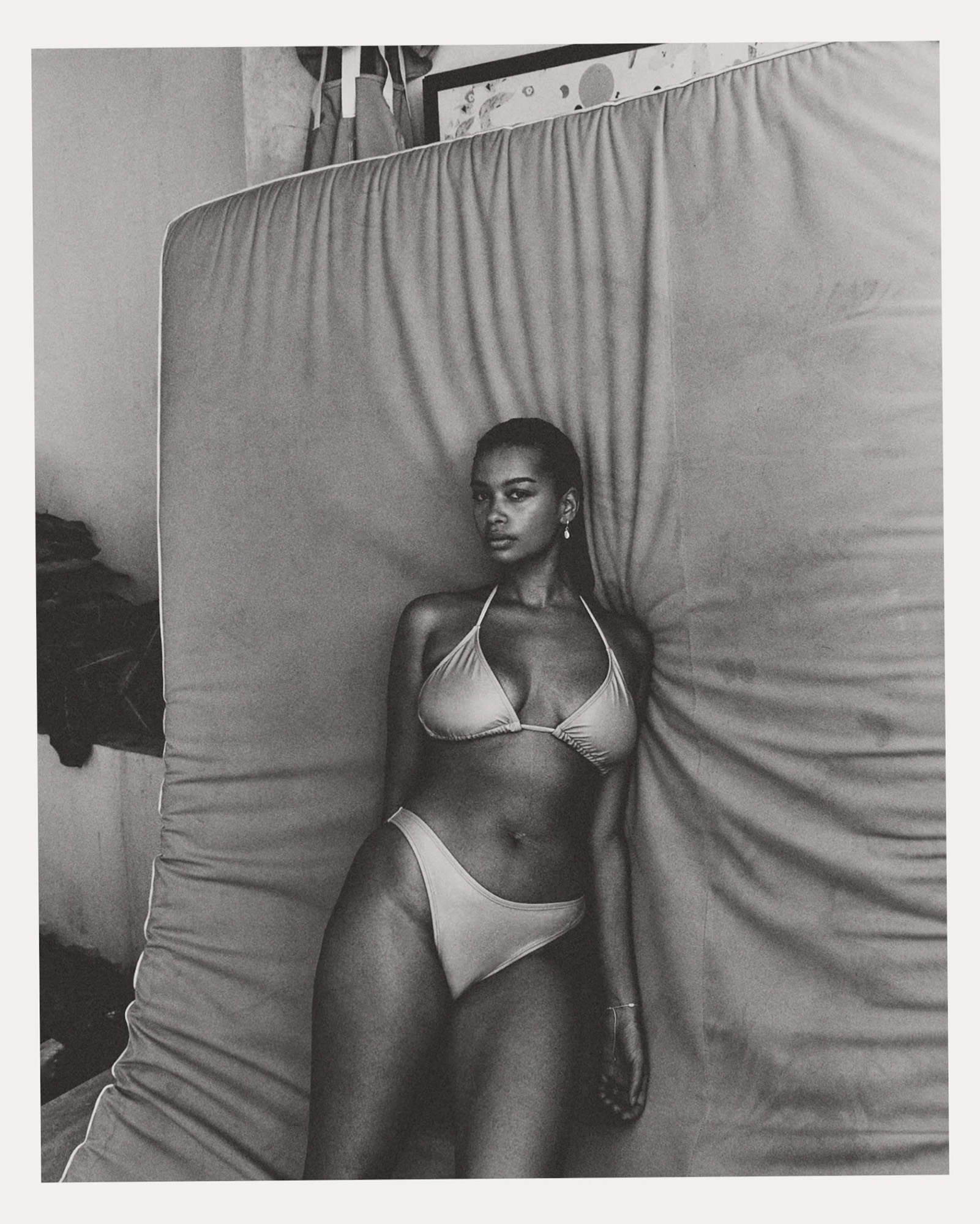 Portrait of a Black woman in a bikini lying on a bed in a neutral indoor setting with diffused lighting, showcasing natural beauty and form.