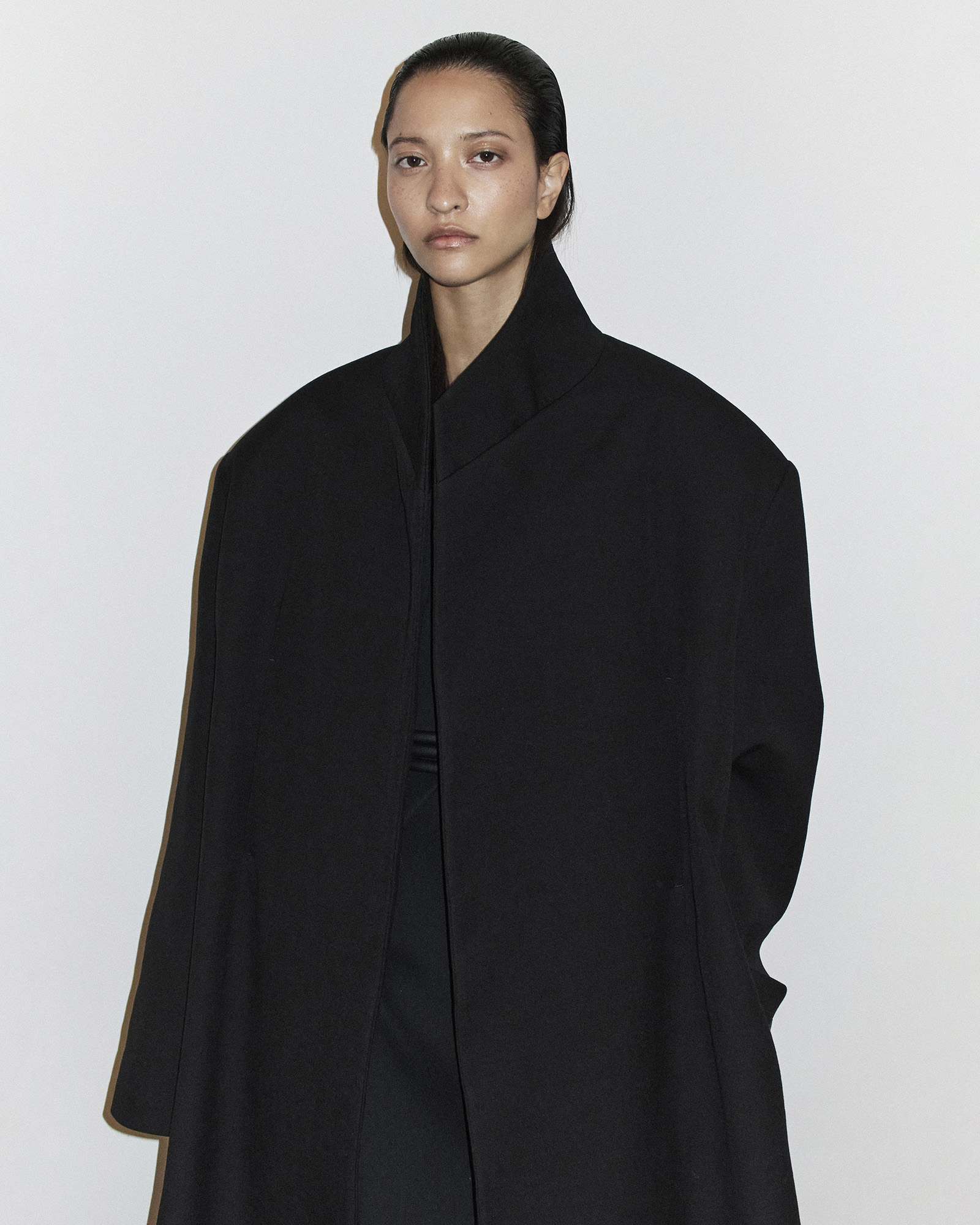 A fashion portrait features a slender adult female in a striking black coat, set against a stark white backdrop in a photo studio.
