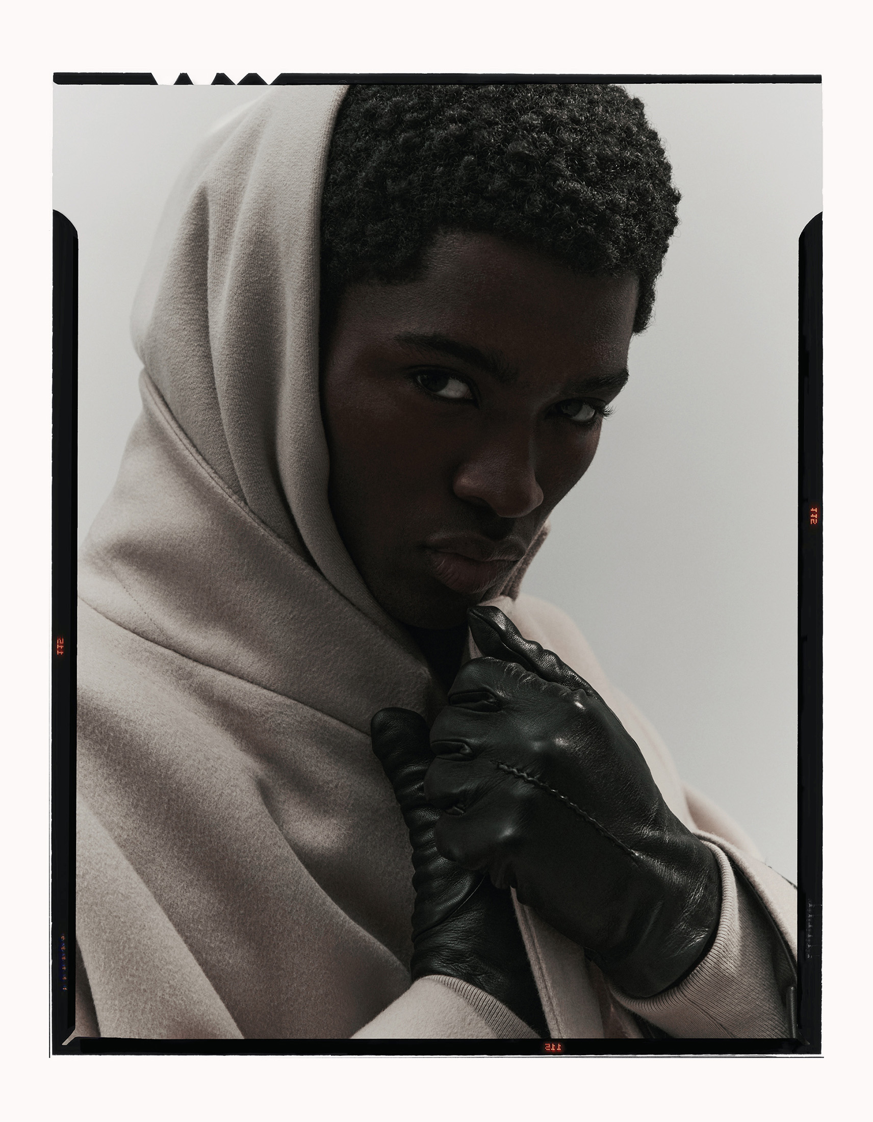 Portrait of an African-American man with dark hair wearing a light beige hooded coat and black leather gloves posing in front of a white background.