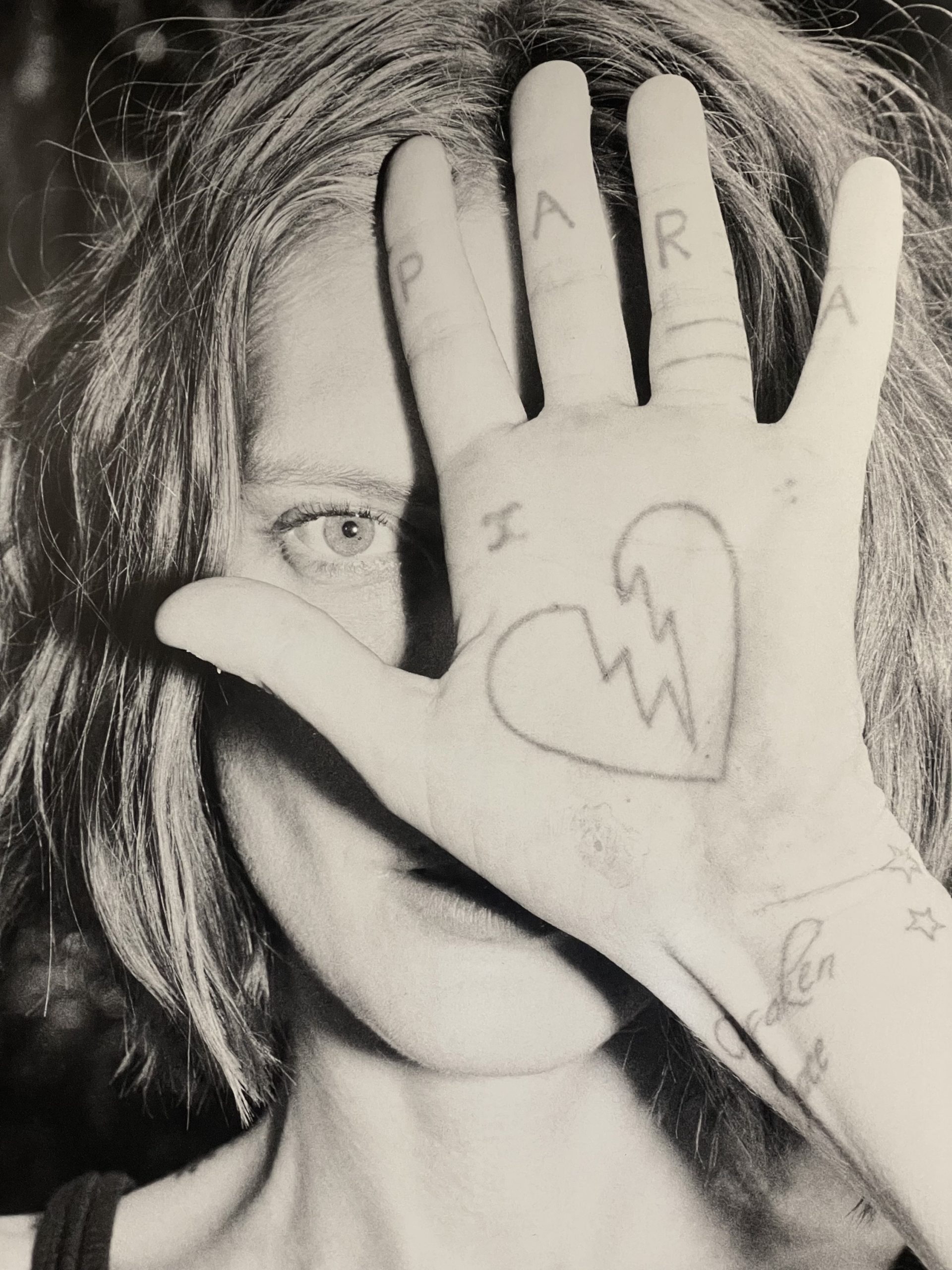Monochrome portrait of a young adult female with a hand covering part of her face, displaying a drawing of a broken heart.