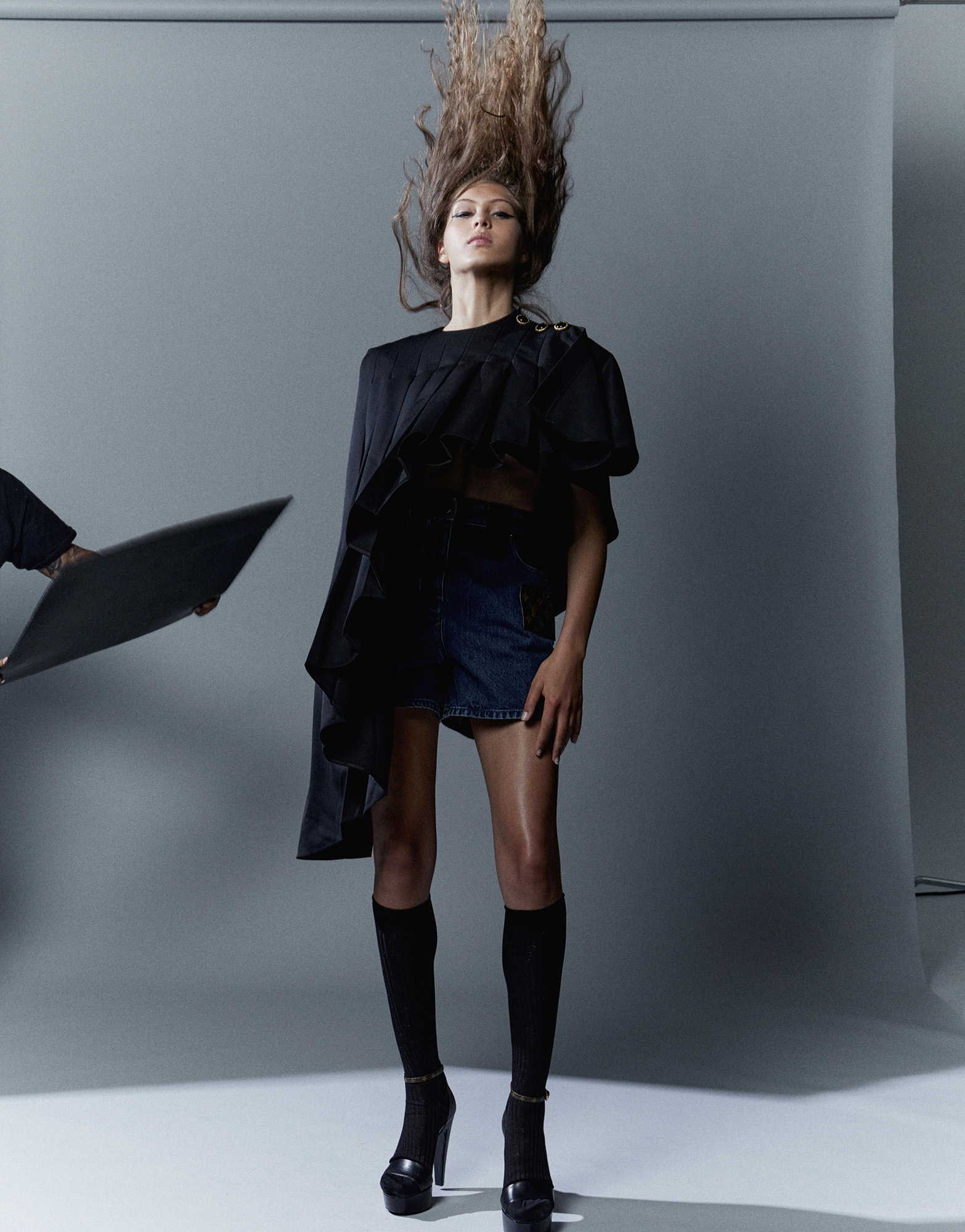 Fashion photo of a woman with long brown hair flying upwards, wearing a black top, short denim skirt, knee-high socks, and high heels in studio.