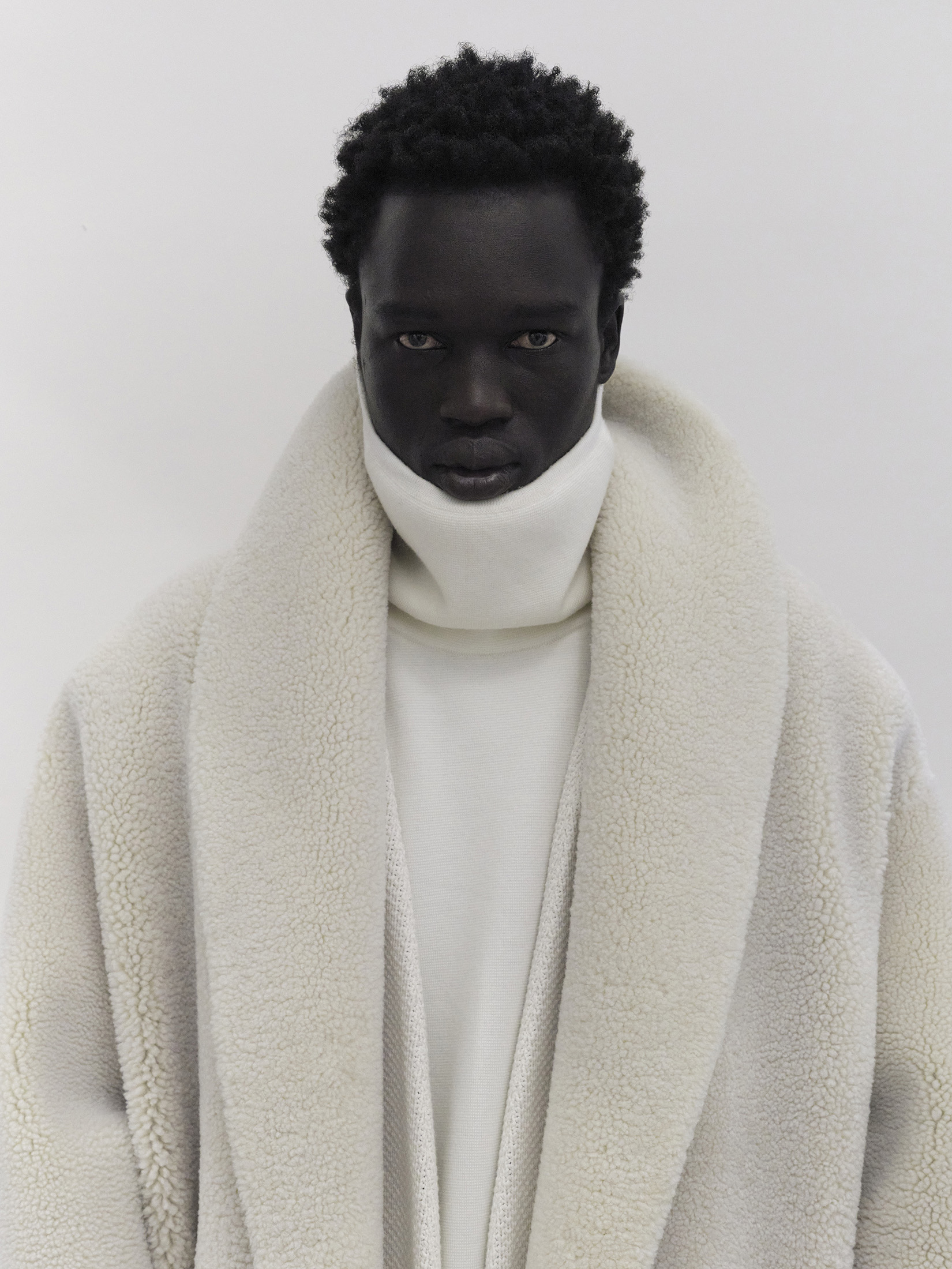Portrait of a dark-skinned male model in a white turtleneck and cream sherpa coat, posing against a white backdrop, studio lighting.