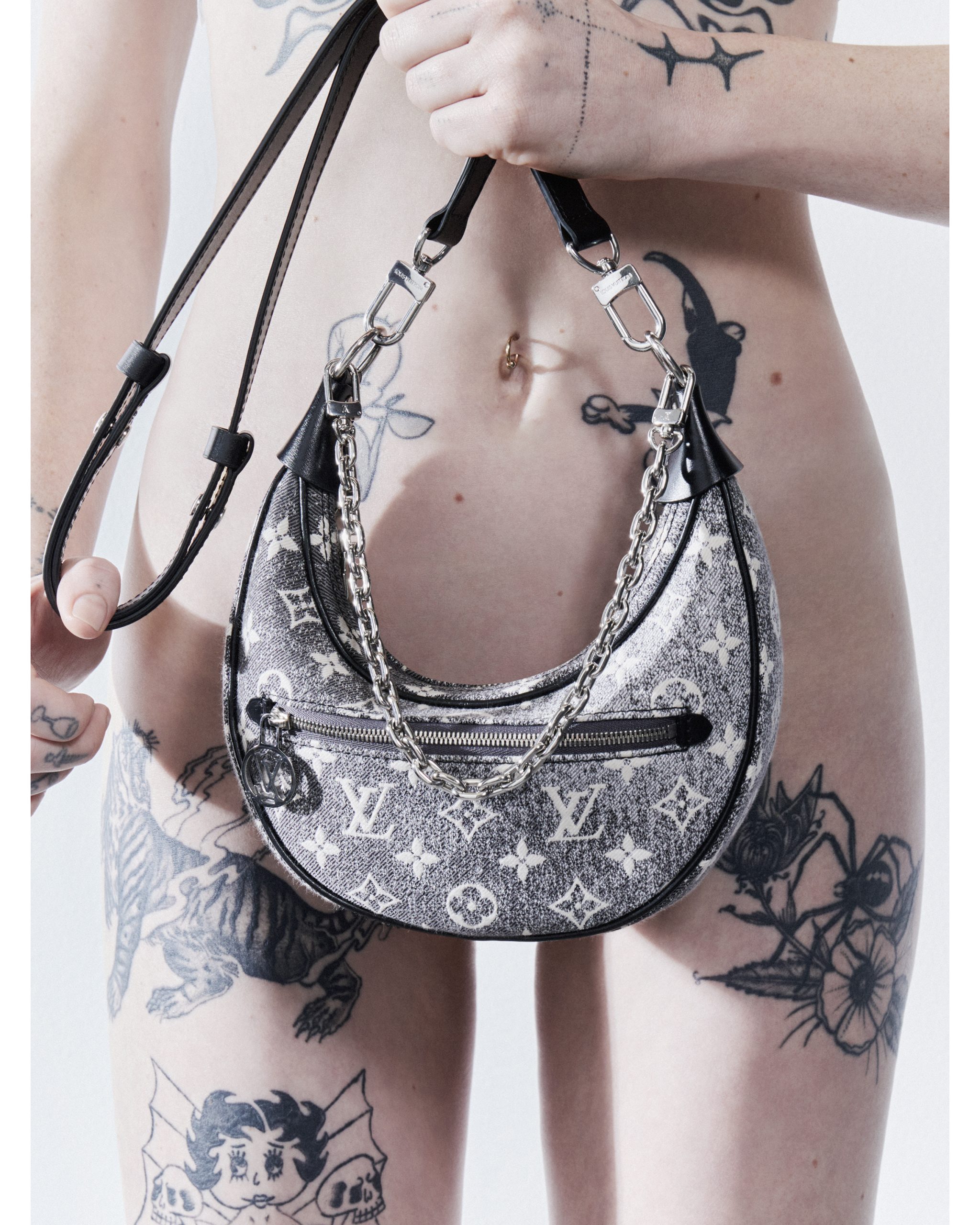 Tattooed torso of model with branded handbag. Half-moon shaped, neutral color with black trim and silver hardware on white backdrop.