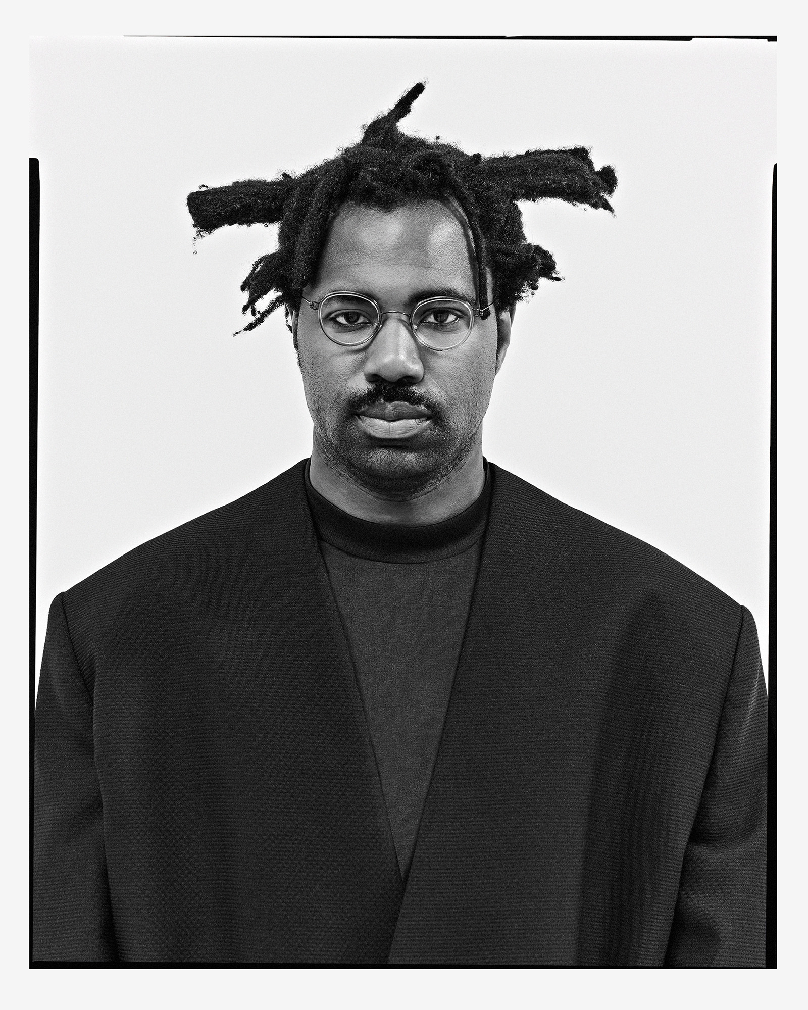 Portrait of an adult male with dreadlocks wearing glasses and a black blazer in a photo studio with a white background, close-up.