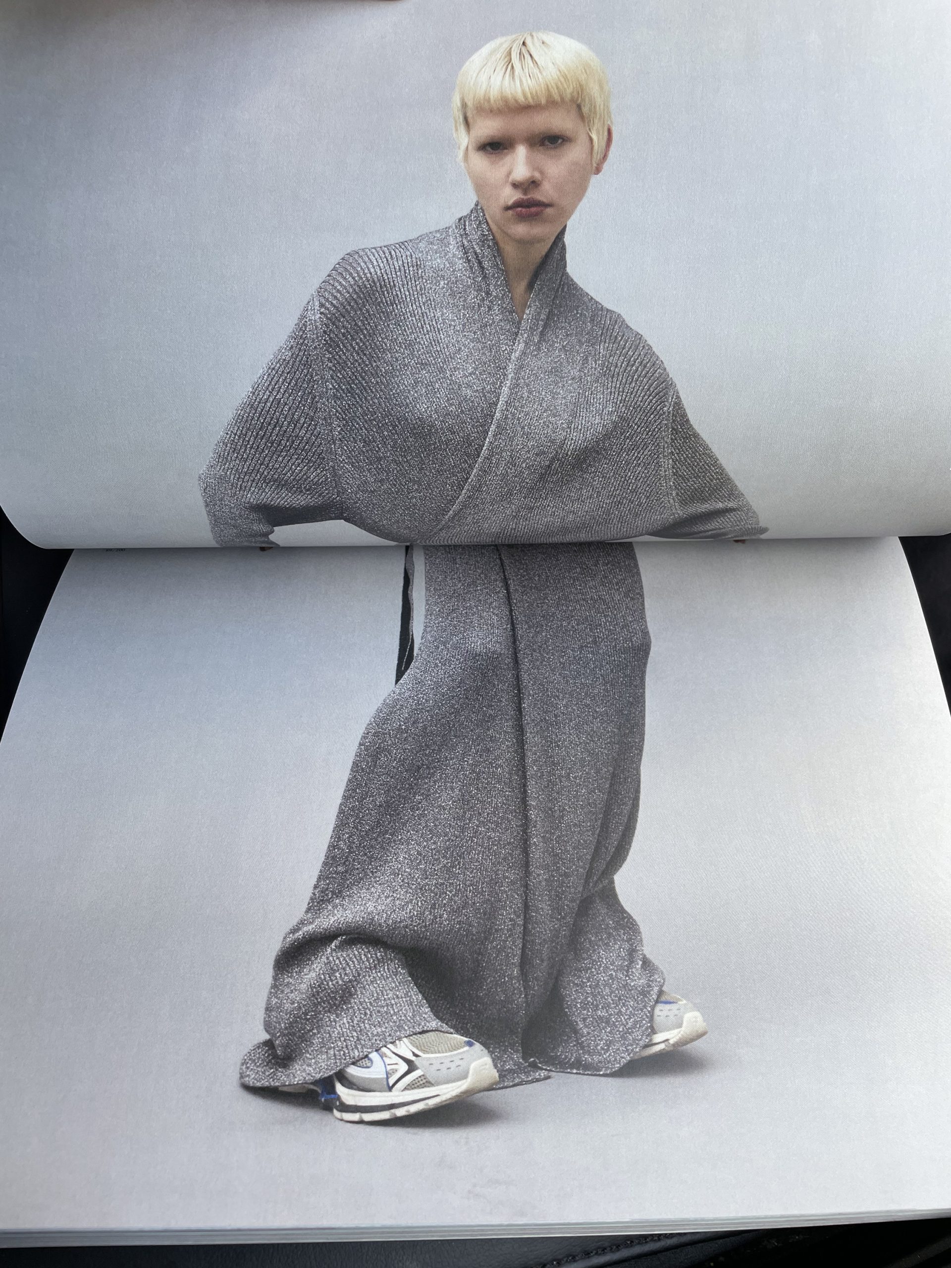 Fashion editorial featuring a blonde model wearing a shimmering grey wrap-style dress and sneakers, posed on a neutral grey backdrop.