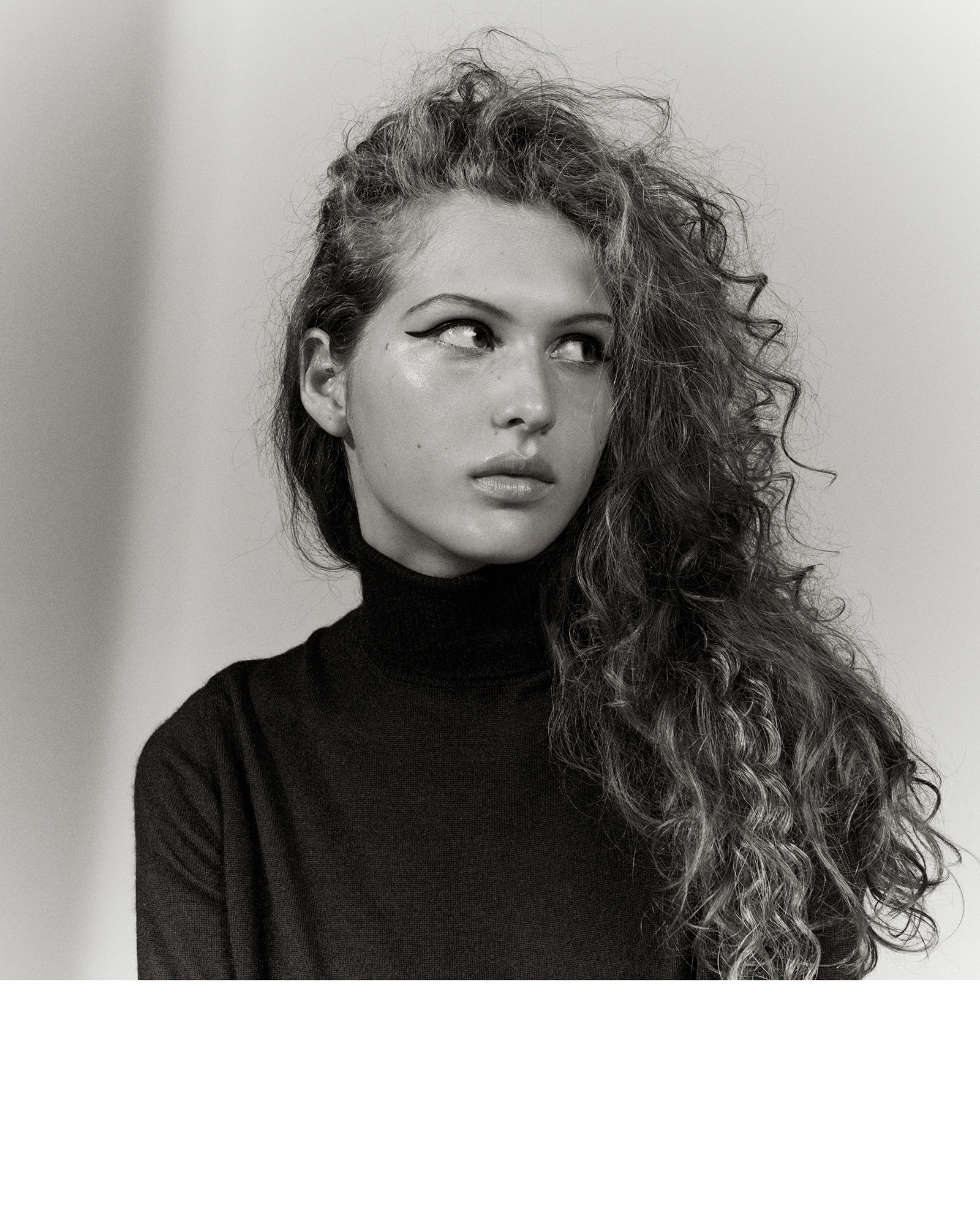 Monochrome portrait of a young adult woman with long curly hair wearing a turtleneck sweater and dark eyeliner, looking off to the side.