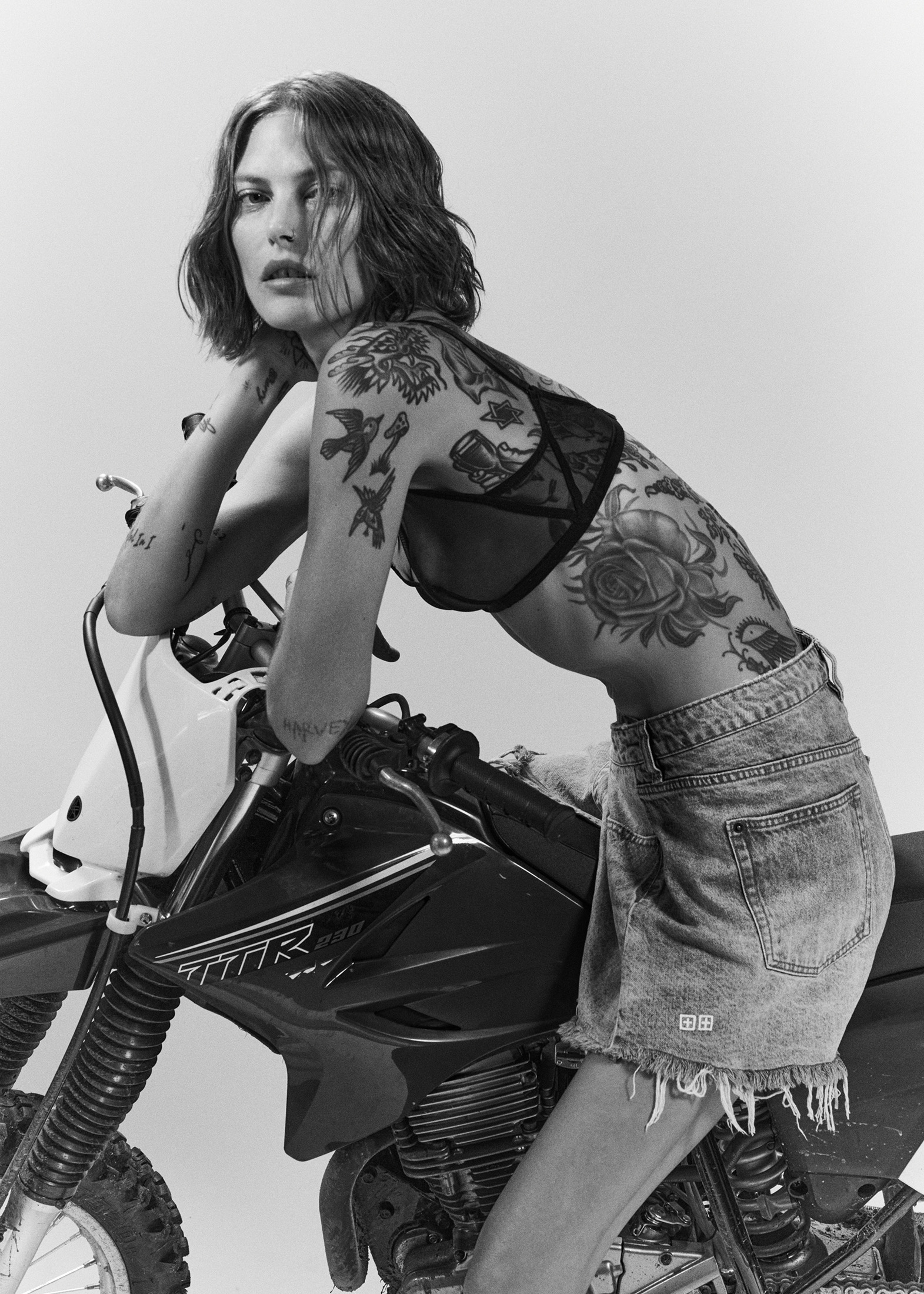Monochrome portrait of tattooed woman in bra and denim skirt posing on a black and white dirt bike in a minimalist photo studio.