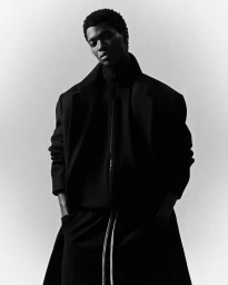Monochrome portrait of a black man wearing a dark structured coat with his hands in his pockets against a white gradient background.