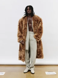 Black man wearing a brown fur coat over light-colored trousers against a plain white studio backdrop.