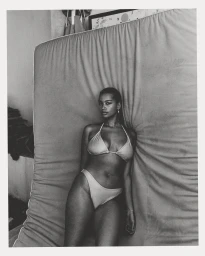 Black and white portrait of a woman wearing a bikini, leaning against a large textured mattress in Sydney by Pierre Toussaint.