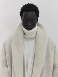 Black man wearing a heavy white shearling coat and high turtleneck collar against a neutral background