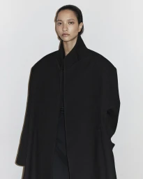 Model with slicked-back hair wearing an oversized black coat against a plain off-white wall background