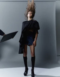 Model with flying long hair wearing a black top and dark shorts against a grey seamless studio backdrop.