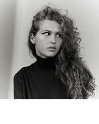 Portrait of a woman with long curly hair and winged eyeliner wearing a black turtleneck sweater against a plain background