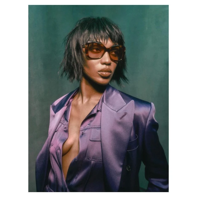 Close-up portrait of an adult woman with short dark hair and sunglasses, wearing an open purple blazer against a green background.