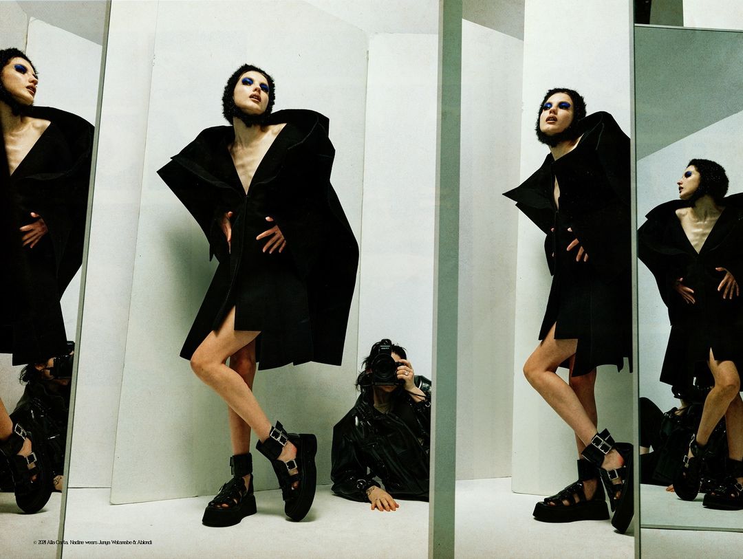 Fashion editorial shot with a slender model in a black outfit posing in front of mirrors with a photographer capturing the scene.